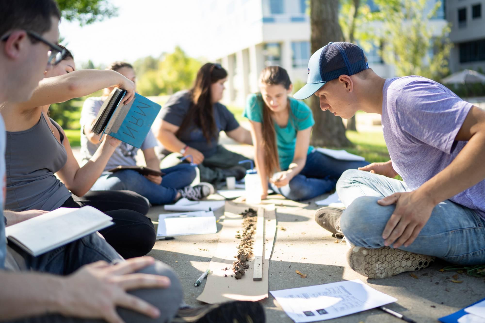 Students sitting on the ground outside gathered around a soil sample and analyzing it